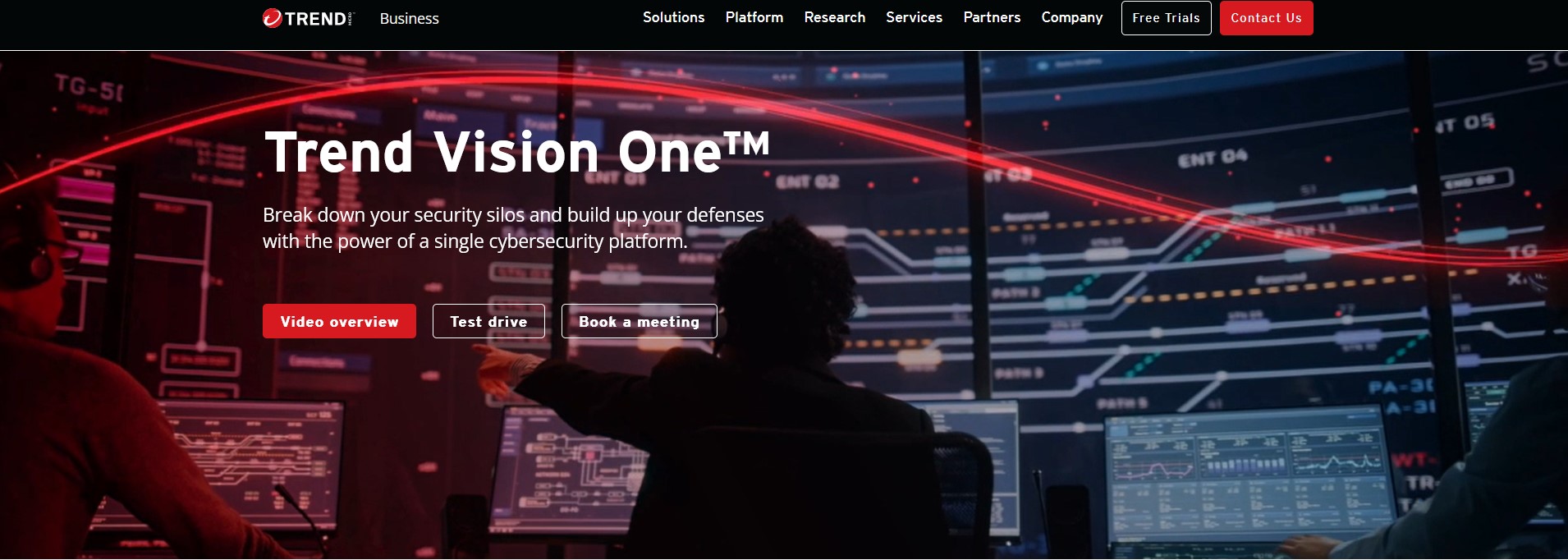 Trend Vision One Image: A security operations center with a person monitoring multiple screens. The text "Trend Vision One™" is displayed with the tagline "Break down your security silos and build up your defenses with the power of a single cybersecurity platform." Buttons for a video overview, test drive, and booking a meeting are included, with a red theme highlighting key elements.